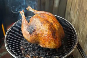 Whole turkey smoking on a charcoal grill with smoke rising, creating a rich golden-brown crust.