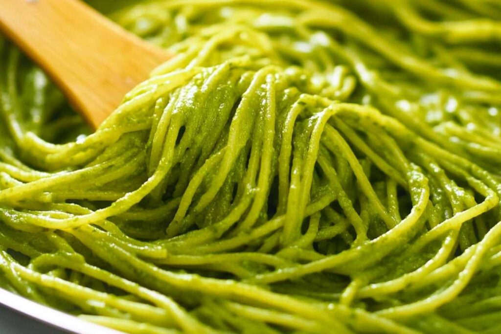 Spaghetti coated in a creamy green pesto sauce, being stirred with a wooden spoon.