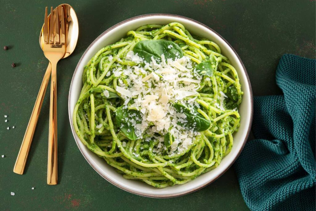 A bowl of green spaghetti garnished with spinach leaves and grated parmesan cheese, served with golden cutlery on a green background.