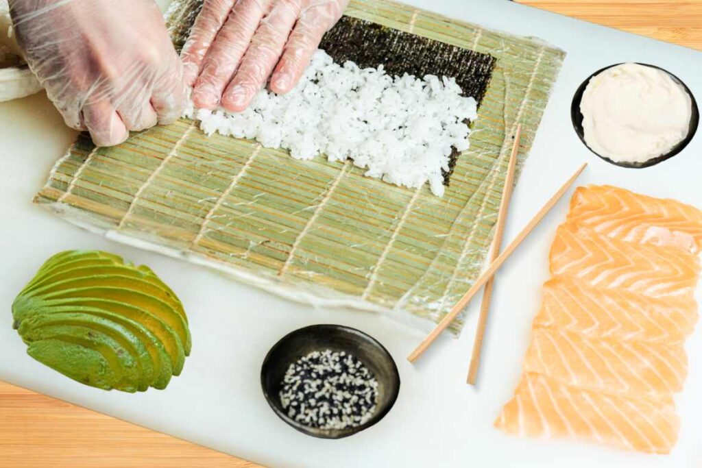 Preparing sushi using a bamboo mat, with ingredients including sushi rice, sliced avocado, fresh salmon, sesame seeds, and cream cheese.