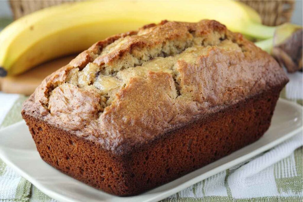 A freshly baked banana bread loaf with a golden crust, resting on a white plate with ripe bananas in the background.