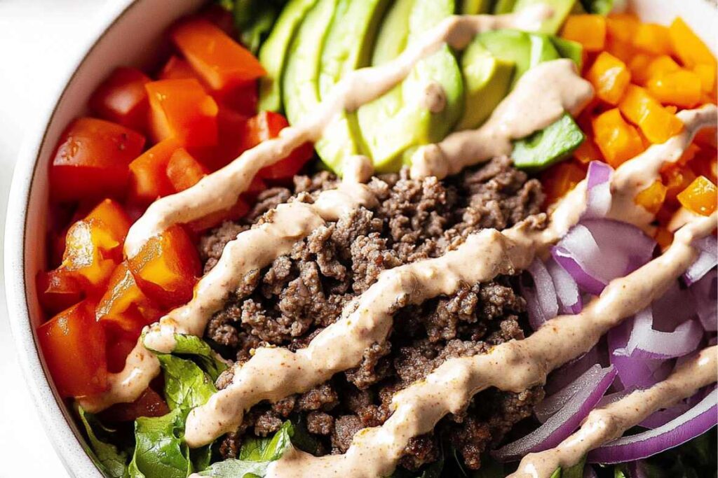 A colorful burger bowl with ground beef, diced tomatoes, avocado slices, red onion, bell peppers, and a drizzle of creamy dressing.