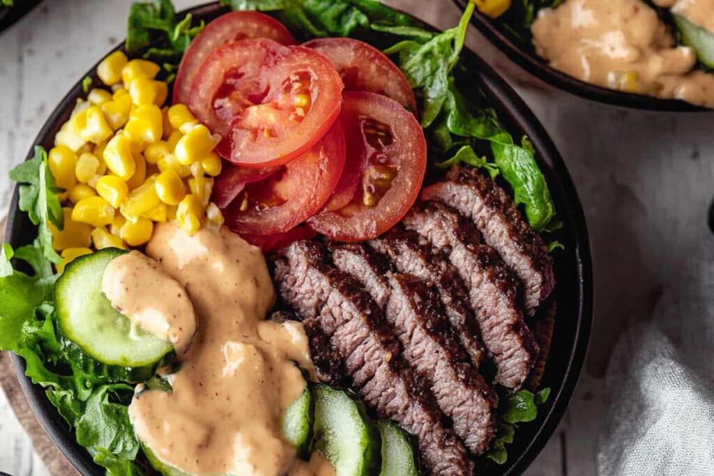 A healthy burger bowl with fresh greens, sliced tomatoes, cucumbers, grilled steak, corn, and creamy dressing.