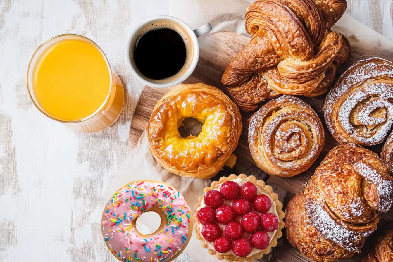 Can You Eat Pastry for Breakfast? A variety of freshly baked pastries, including croissants, cinnamon rolls, danishes, a raspberry tart, and a sprinkled donut, served with orange juice and black coffee on a wooden board.