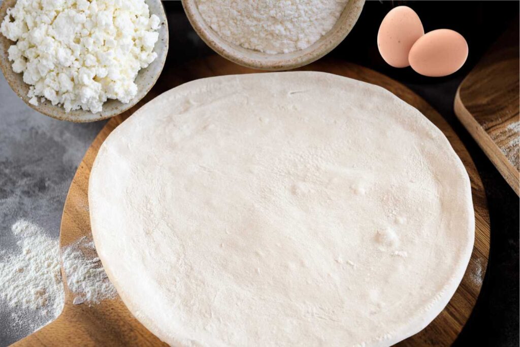 Unbaked cottage cheese pizza crust displayed on a wooden board with bowls of cottage cheese, flour, and eggs in the background.