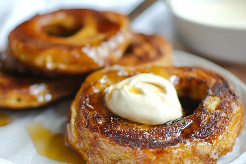 Close-up of a French Toast Bagel topped with butter and drizzled with syrup on a white plate.