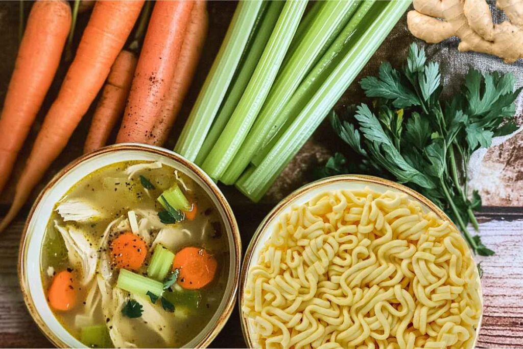 A bowl of ginger chicken soup surrounded by fresh carrots, celery, herbs, and noodles.