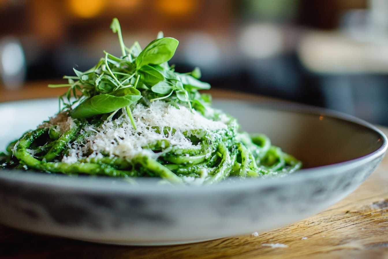 A bowl of green spaghetti topped with grated Parmesan cheese and fresh basil leaves, served on a wooden table in a cozy setting.