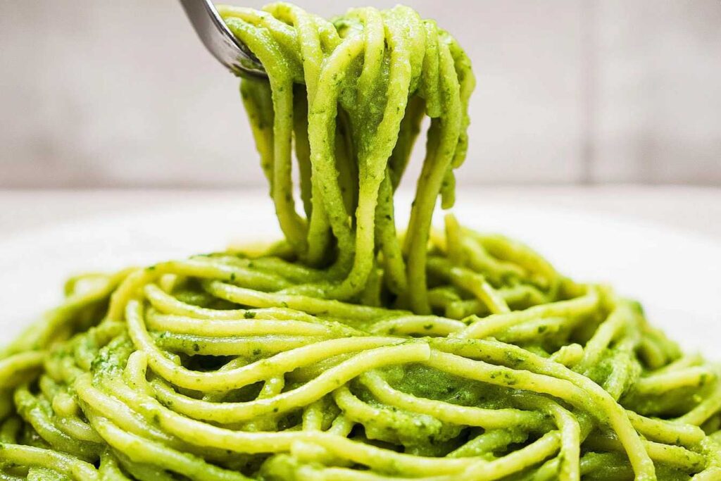 A close-up of green spaghetti being twirled on a fork, coated in creamy pesto sauce, served on a white plate.