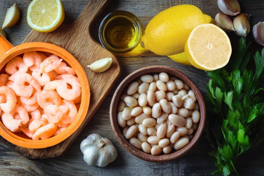 Fresh ingredients for lemony shrimp and bean stew including shrimp, white beans, lemons, garlic cloves, olive oil, and parsley on a rustic wooden surface.