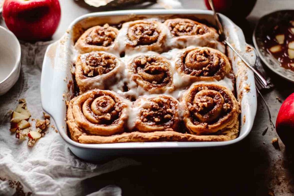 Cinnamon rolls filled with apple pie filling, topped with icing and served in a white baking dish.