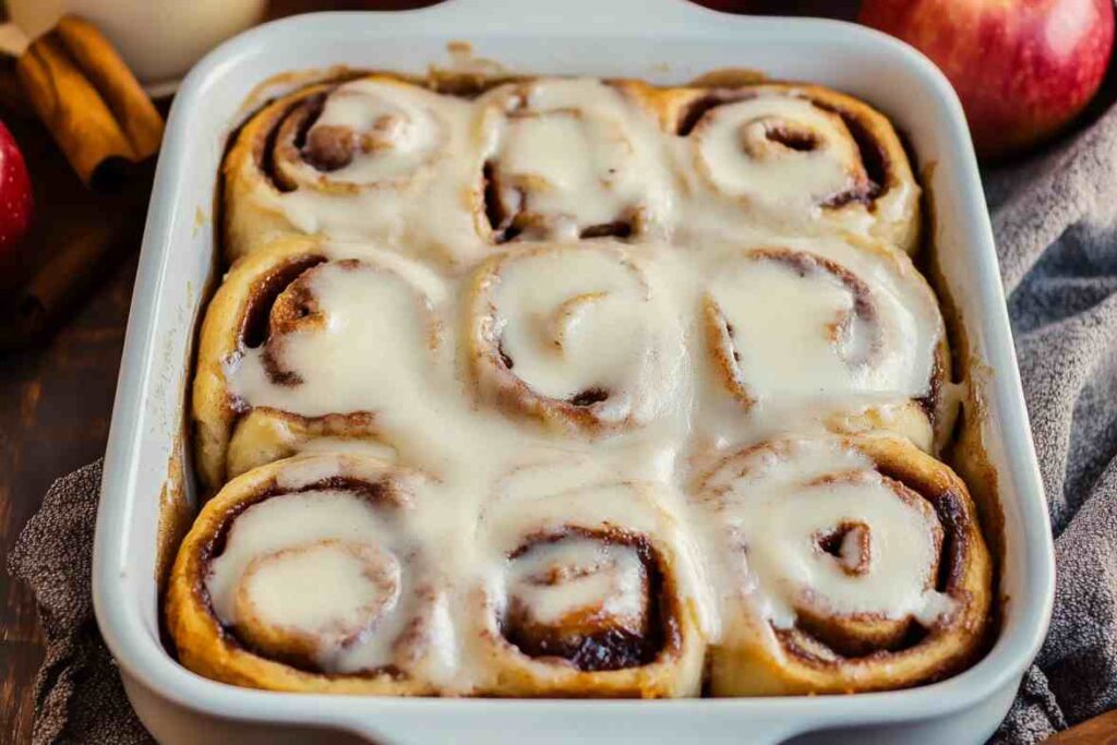 Freshly baked cinnamon rolls filled with apple pie filling, topped with creamy icing in a white baking dish.