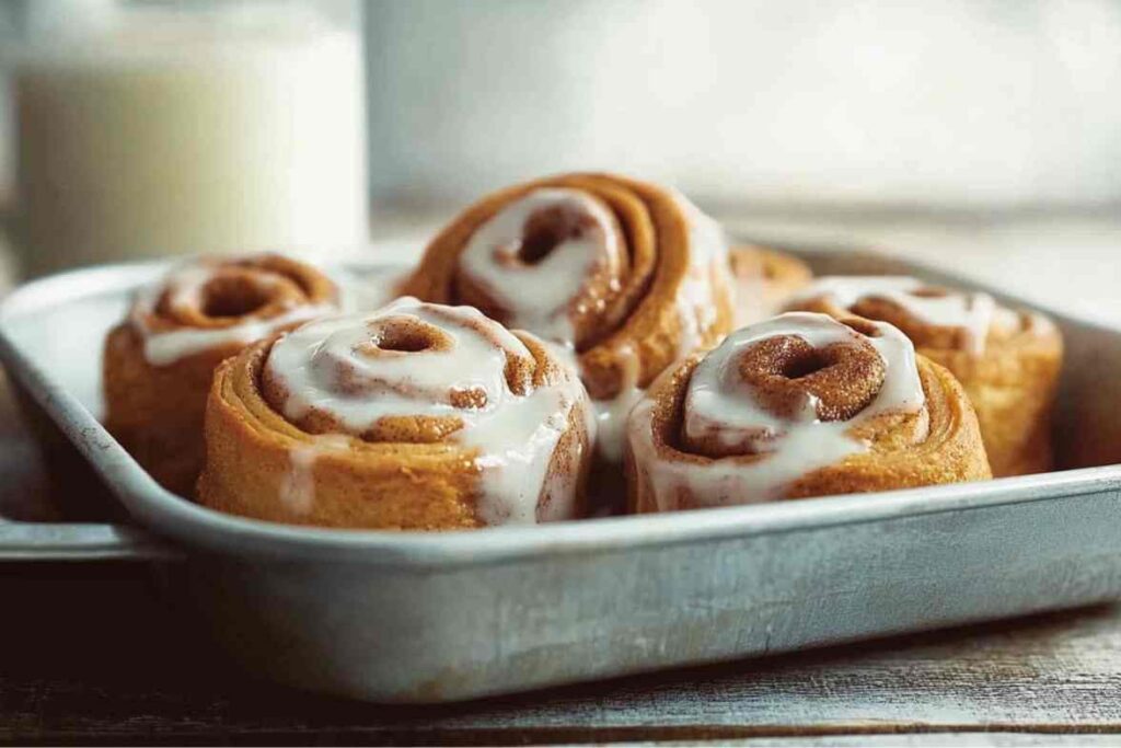 A close-up of freshly baked cinnamon rolls in a metal baking tray, topped with a smooth icing glaze.