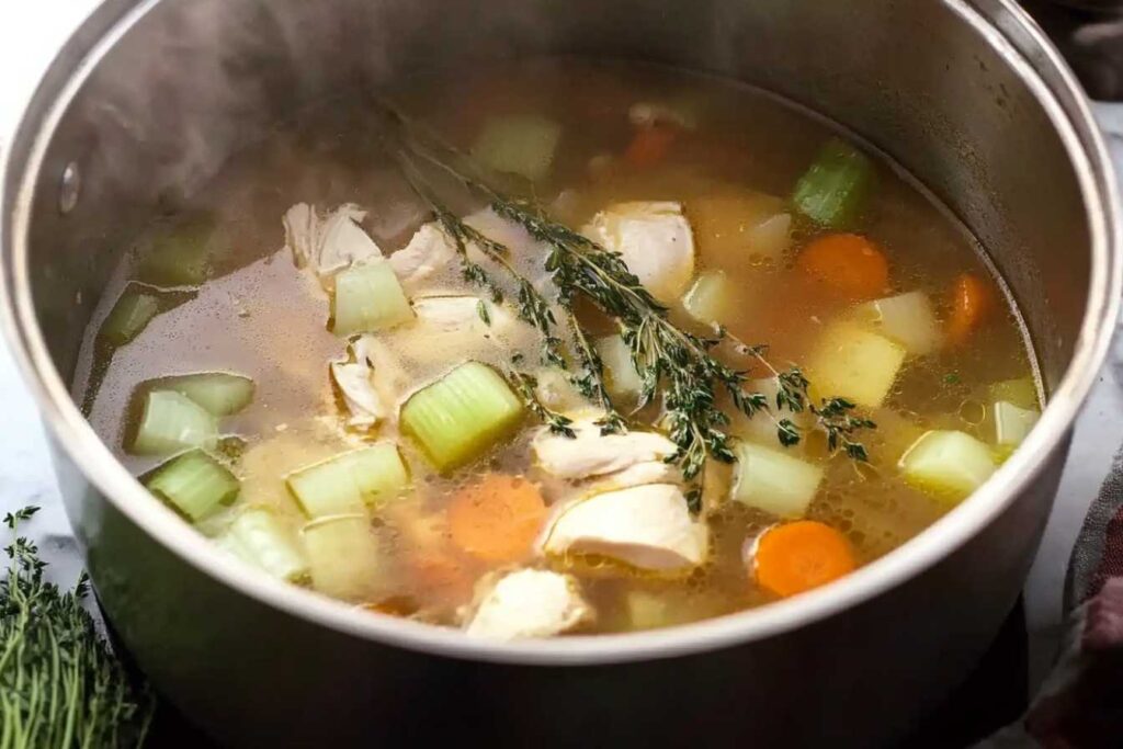 Close-up of Grandma's chicken soup cooking in a pot with carrots, celery, chicken, and fresh thyme.