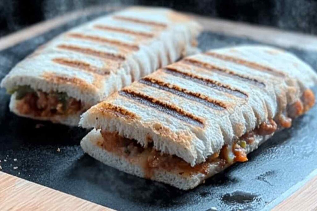 Grilled panini with golden grill marks, filled with a savory mixture, sliced and served on a black slate board.