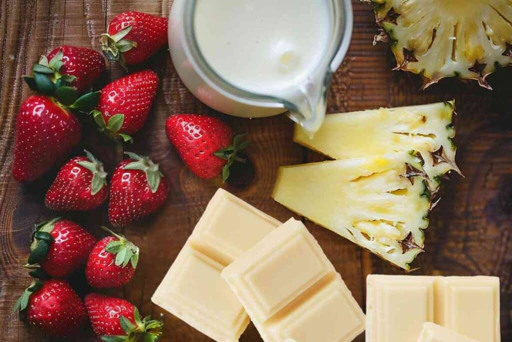 A top-down view of fresh Bahama Mama Tropical Smoothie ingredients, including ripe strawberries, juicy pineapple slices, white chocolate, and creamy coconut milk on a rustic wooden surface.