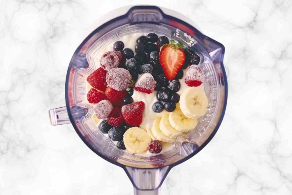 A top-down view of a blender filled with strawberries, blueberries, banana slices, and yogurt, ready to be blended into a Strawberry Blueberry Banana Smoothie.