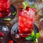 A vibrant cranberry mocktail in a tall glass filled with ice, garnished with fresh cranberries and mint leaves, served on a rustic background with scattered cranberries and ice cubes.