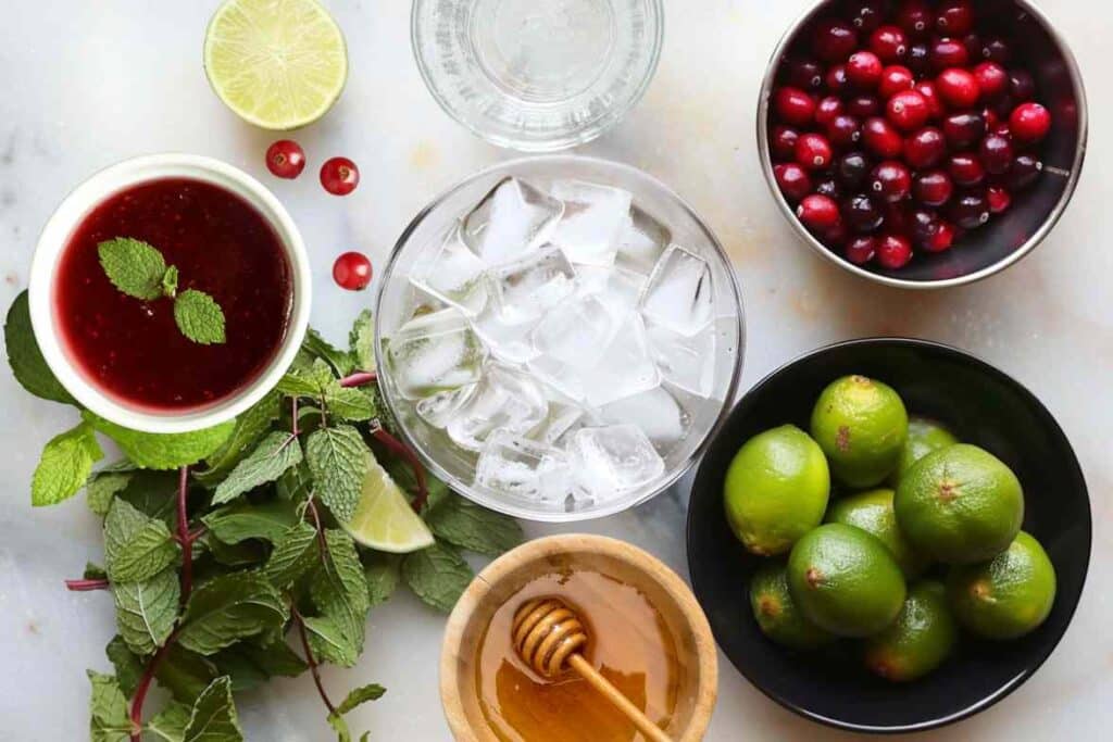 A top-down view of fresh ingredients for a cranberry mocktail, including cranberries, limes, mint leaves, honey, cranberry juice, ice cubes, and sparkling water, arranged on a white marble surface.