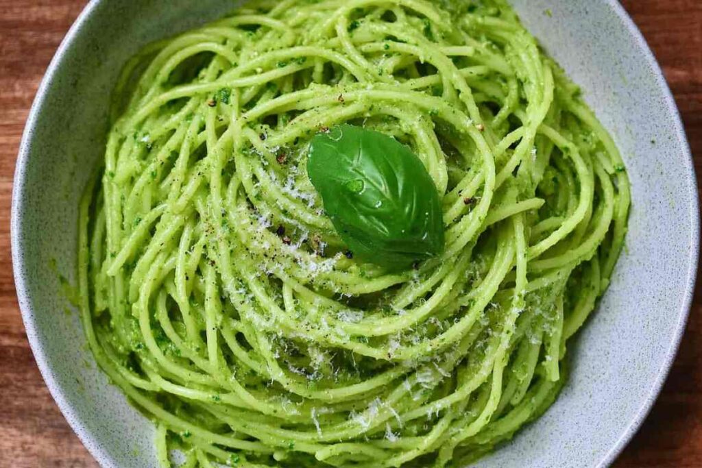 A bowl of green spaghetti garnished with fresh basil and parmesan, showcasing a vibrant and creamy pesto sauce. Learn more about how many calories are in a cup of green spaghetti.