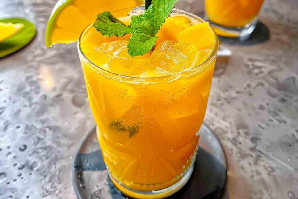 A vibrant mango mocktail served in a glass with ice cubes, garnished with fresh mint and a mango slice, placed on a stylish table with a citrus touch.