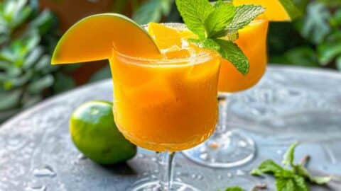 Refreshing mango mocktail served in a glass with ice, garnished with fresh mint and a mango slice, with lime and greenery in the background.