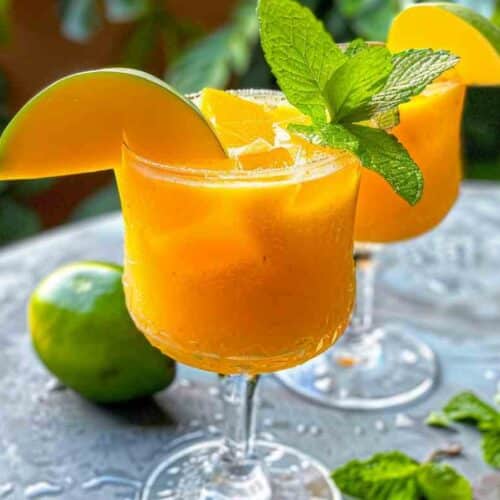 Refreshing mango mocktail served in a glass with ice, garnished with fresh mint and a mango slice, with lime and greenery in the background.