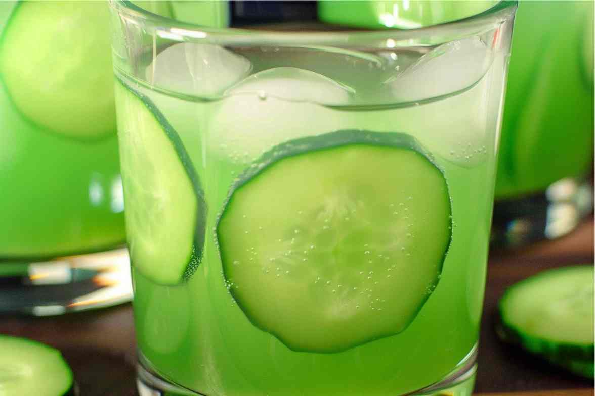 A close-up of a refreshing cucumber mocktail served in a glass filled with ice cubes and thin cucumber slices, with a fizzy texture and a vibrant green hue.