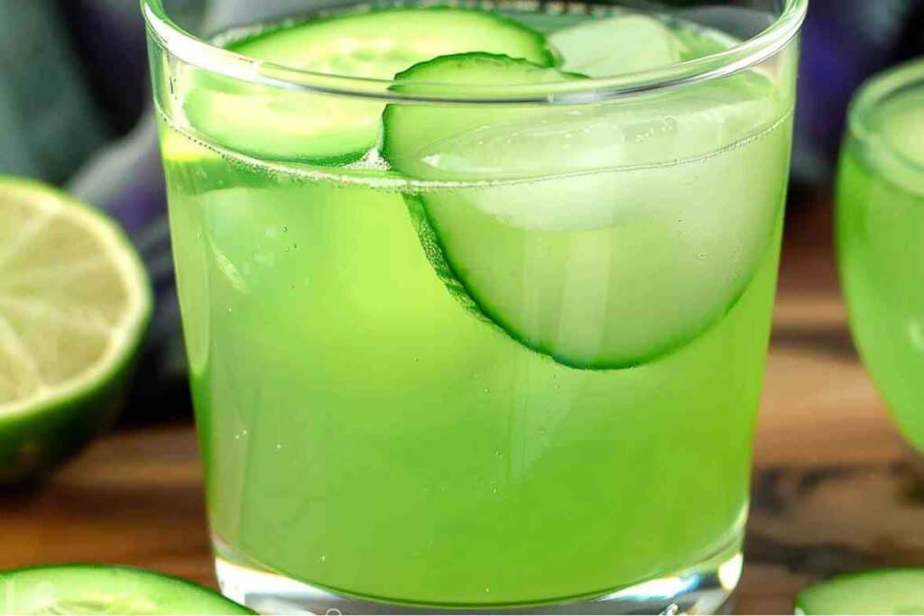 A vibrant green cucumber mocktail served in a clear glass with ice cubes and thin cucumber slices, accompanied by fresh limes in the background.