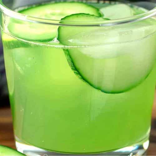 A vibrant green cucumber mocktail served in a clear glass with ice cubes and thin cucumber slices, accompanied by fresh limes in the background.