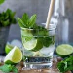 A refreshing mojito mocktail in a glass with fresh mint, lime wedges, ice, and sparkling water, served with a bamboo straw on a wooden surface.