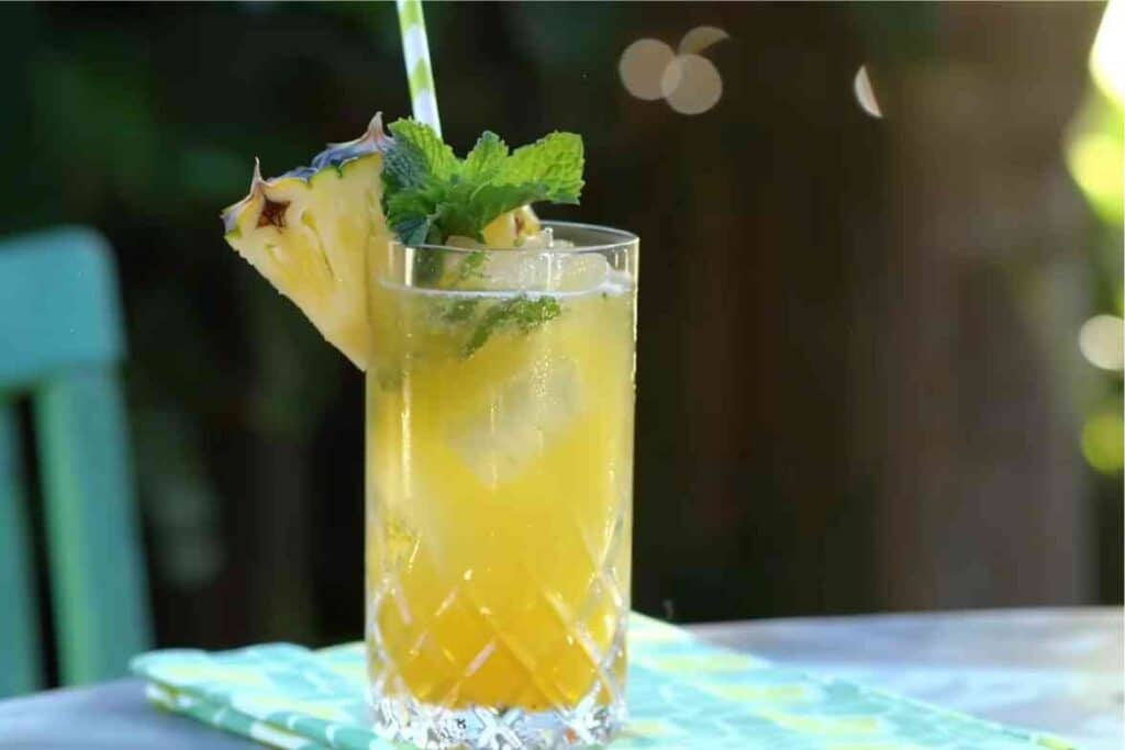 A refreshing pineapple mocktail in a tall, crystal-cut glass, filled with ice, garnished with a fresh pineapple wedge and mint, served with a striped straw on a table with a green outdoor backdrop.